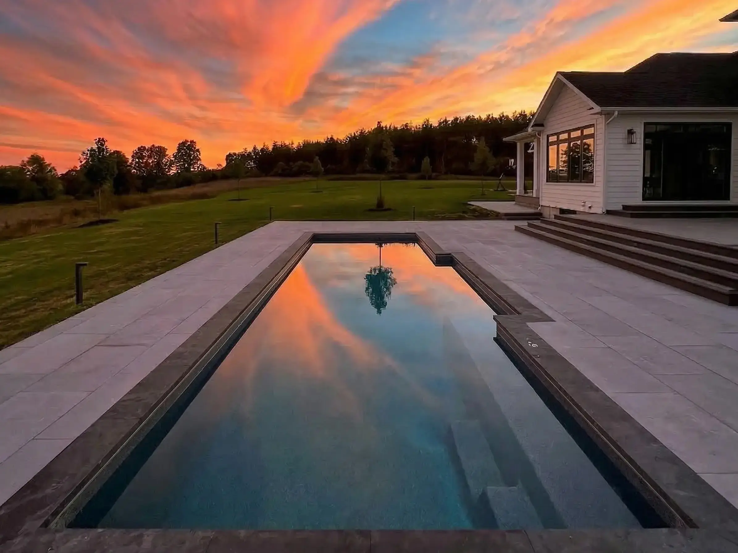 Serene rectangular swimming pool reflecting sunset clouds next to a white farmhouse with dark trim and lawn.