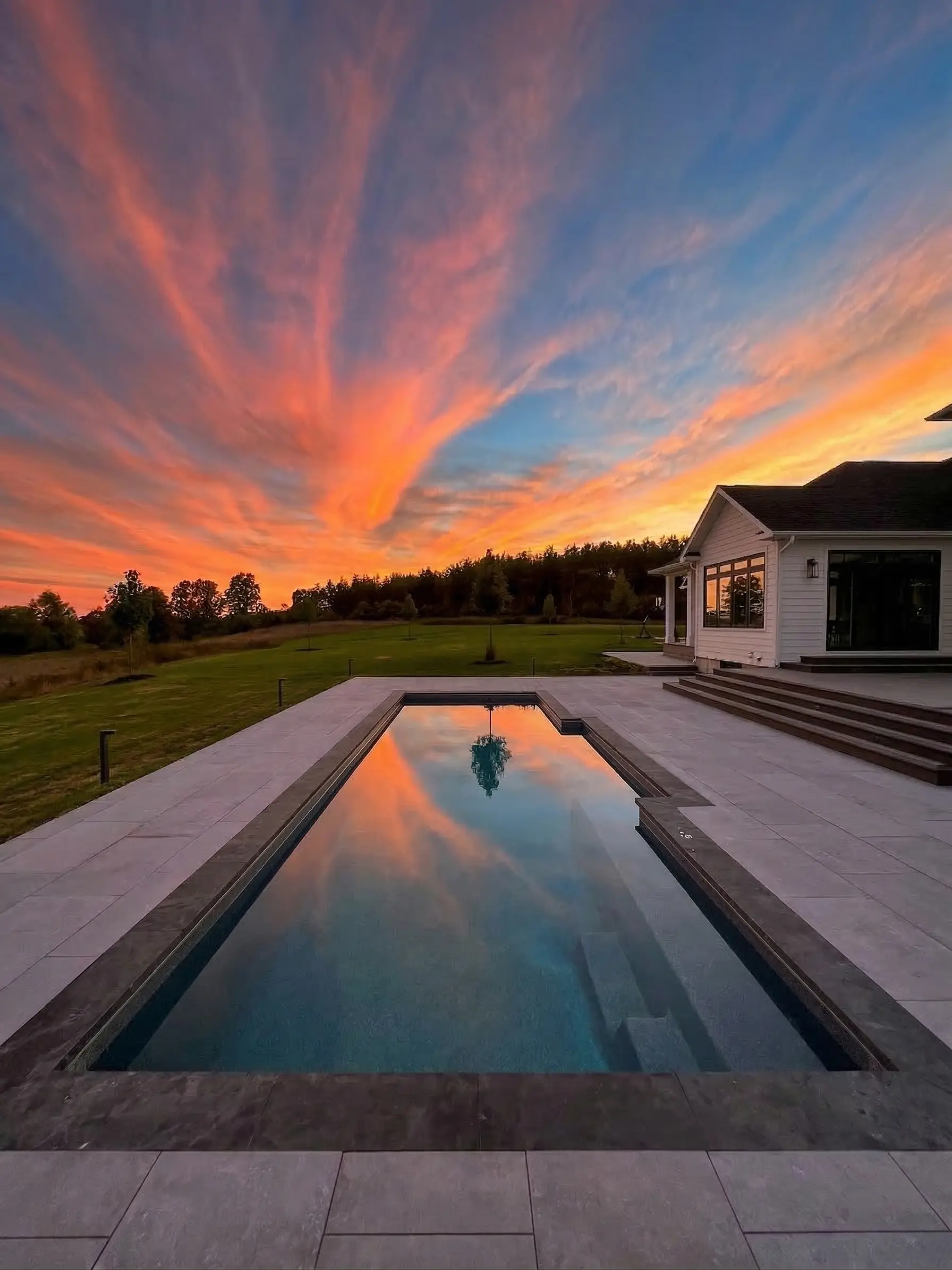 A serene rectangular swimming pool reflecting brilliant orange and pink sunset clouds, located next to a white farmhouse with dark trim and a large grassy lawn.