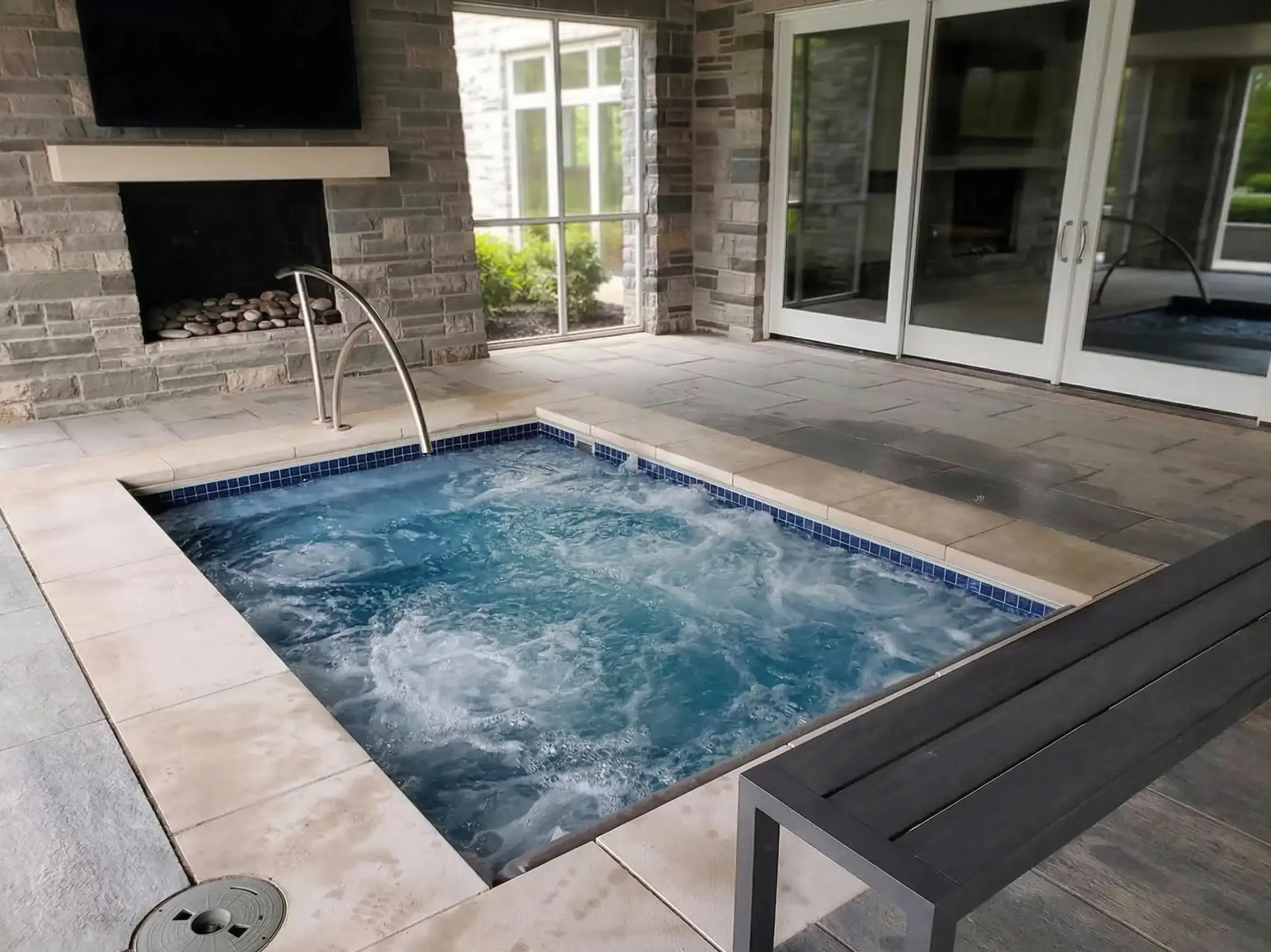 A square built-in hot tub with active jets and a stainless steel handrail, located in a covered stone patio room featuring a fireplace, a gray bench, and large sliding glass doors.