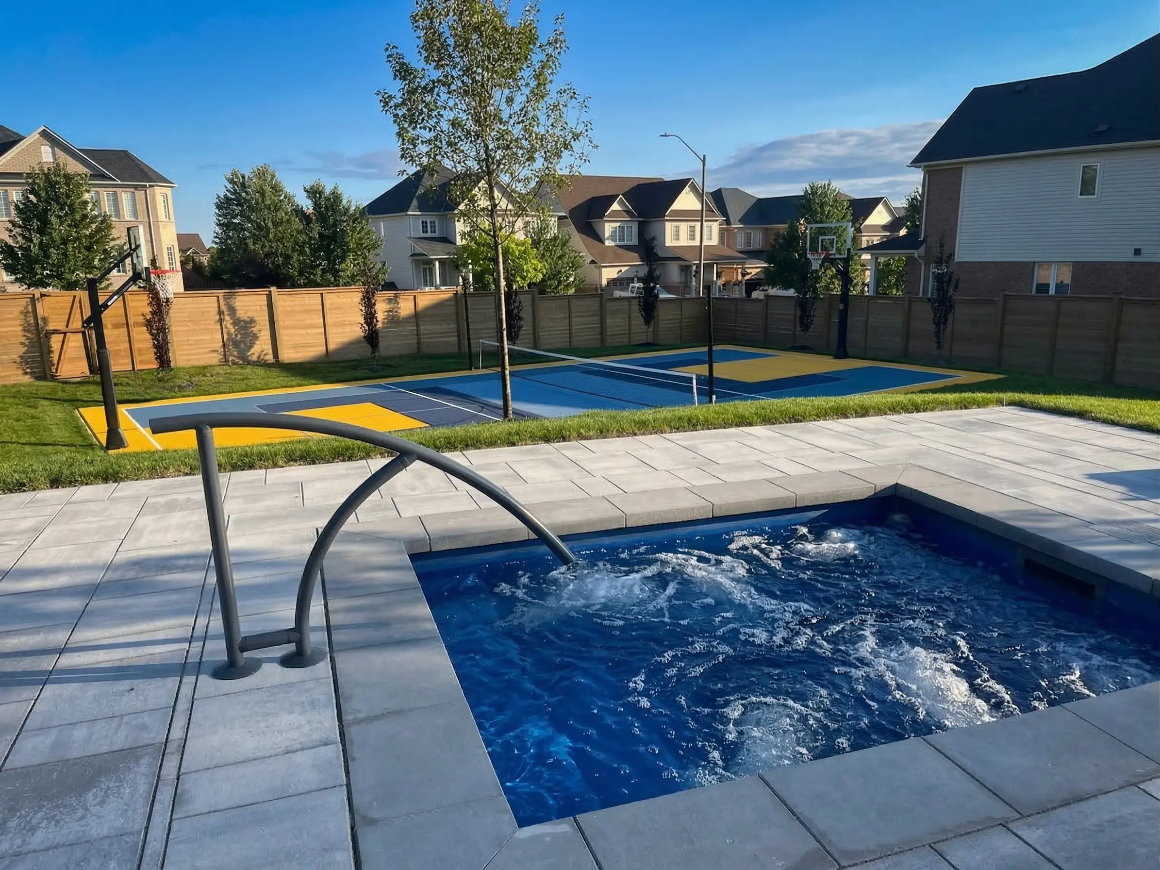 A rectangular hot tub with bubbling water and a metal handrail set into a gray stone patio, looking out over a grassy yard and a colorful multi-sport court.