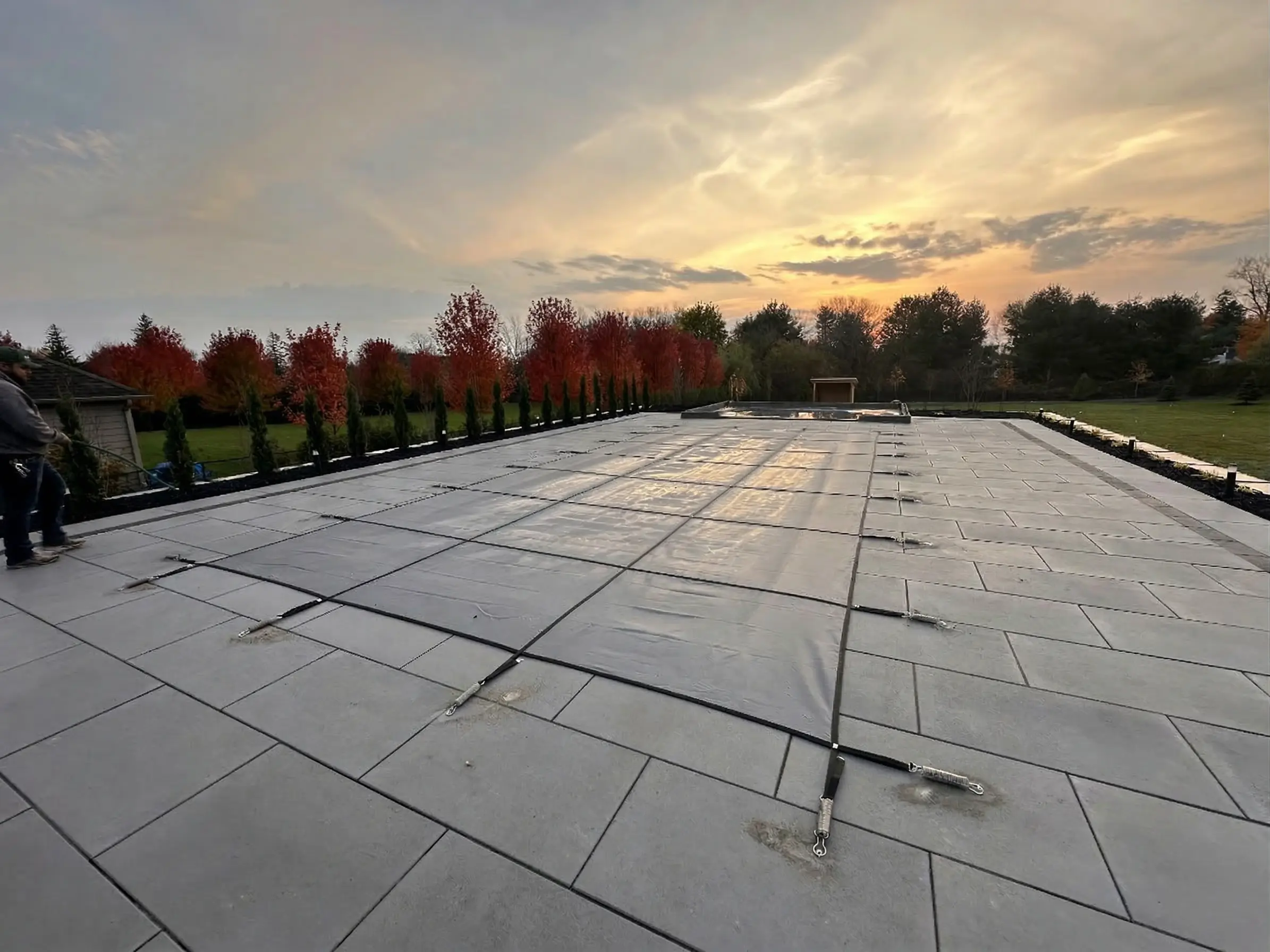A large rectangular swimming pool secured for the season with a gray safety cover anchored into a stone patio, set against a backdrop of red autumn trees and a glowing sunset sky.