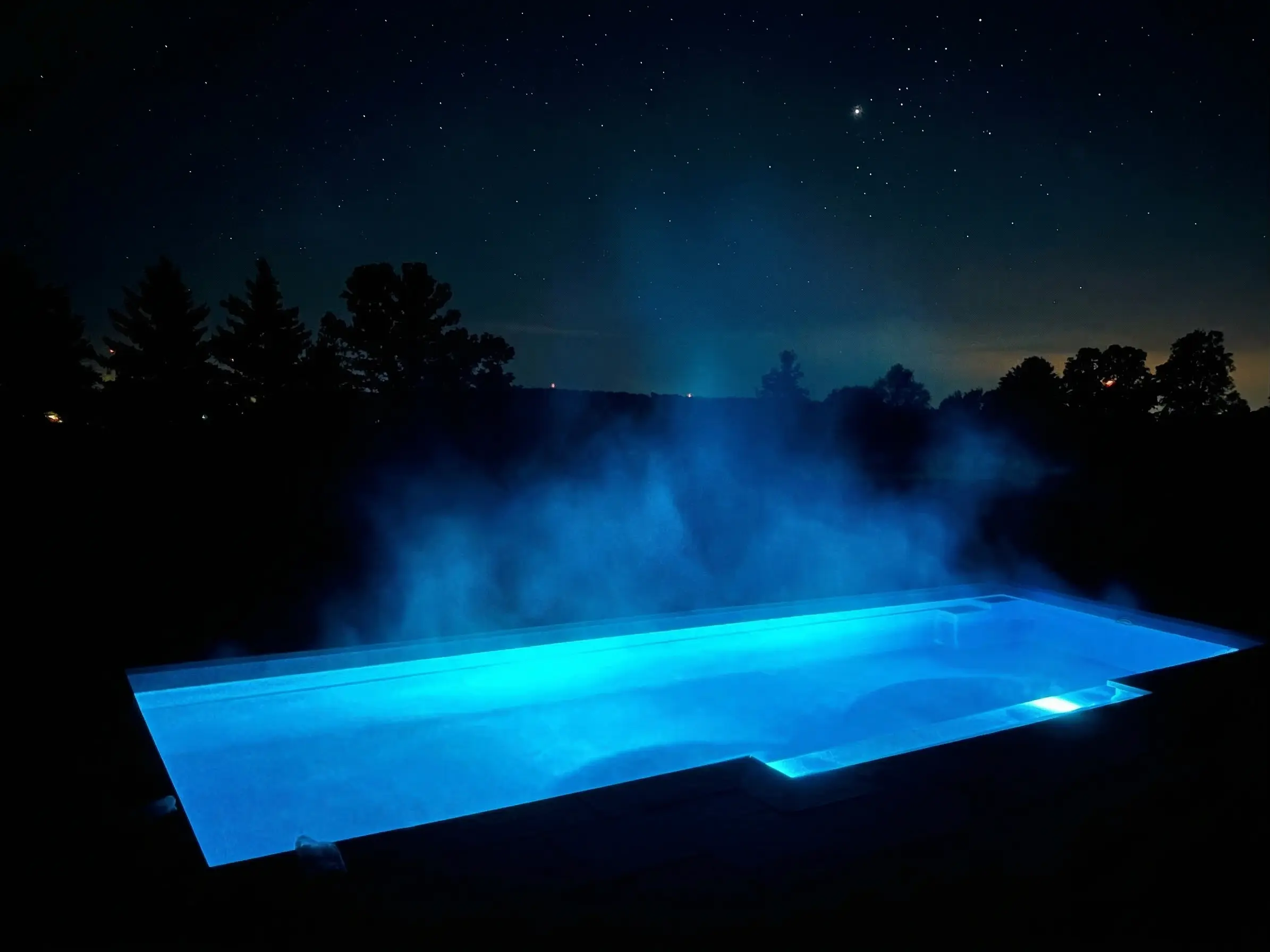 A swimming pool illuminated by intense blue underwater lights at night, with thick steam rising from the warm water against a dark silhouette of trees and a starry sky.