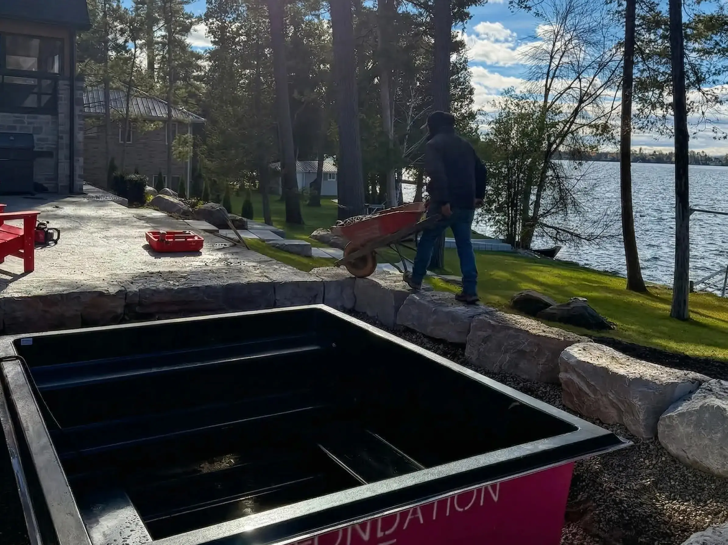 A landscaping worker pushes a wheelbarrow past a black fiberglass plunge pool waiting to be installed in a stone patio, overlooking a scenic lake surrounded by tall pine trees.